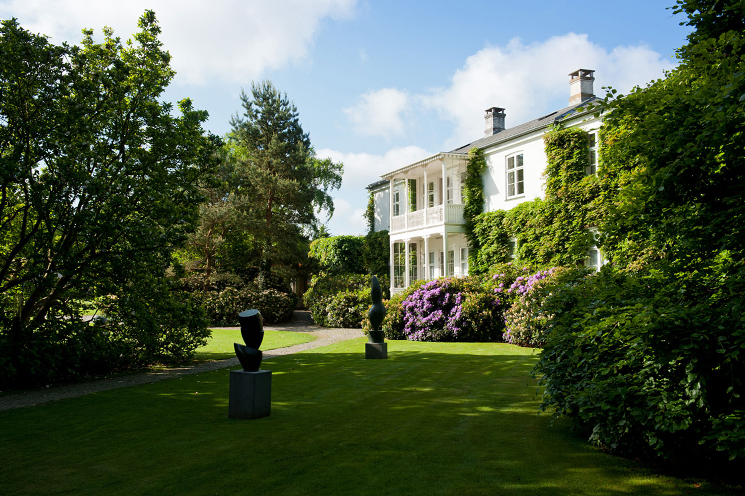 Two sculptures on a grassy area, framed by trees with a building peeking through.