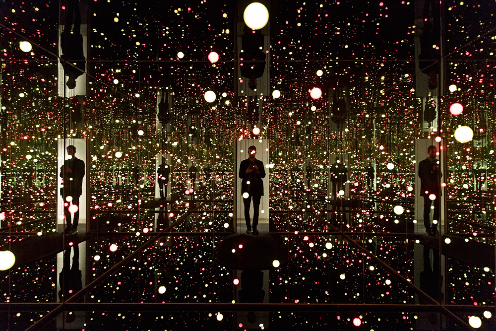 Man standing in a dark mirrored room filled with numerous small lights.