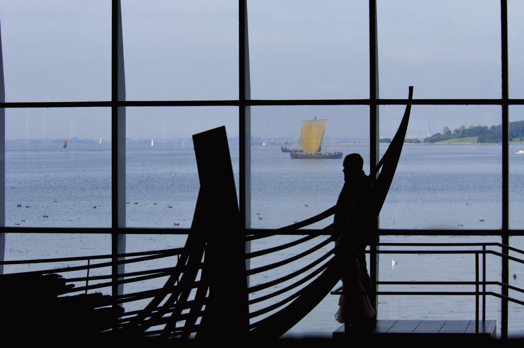 Visitor silhouetted inside, viewing Viking ship skeletons while full ships are seen through the museum windows.