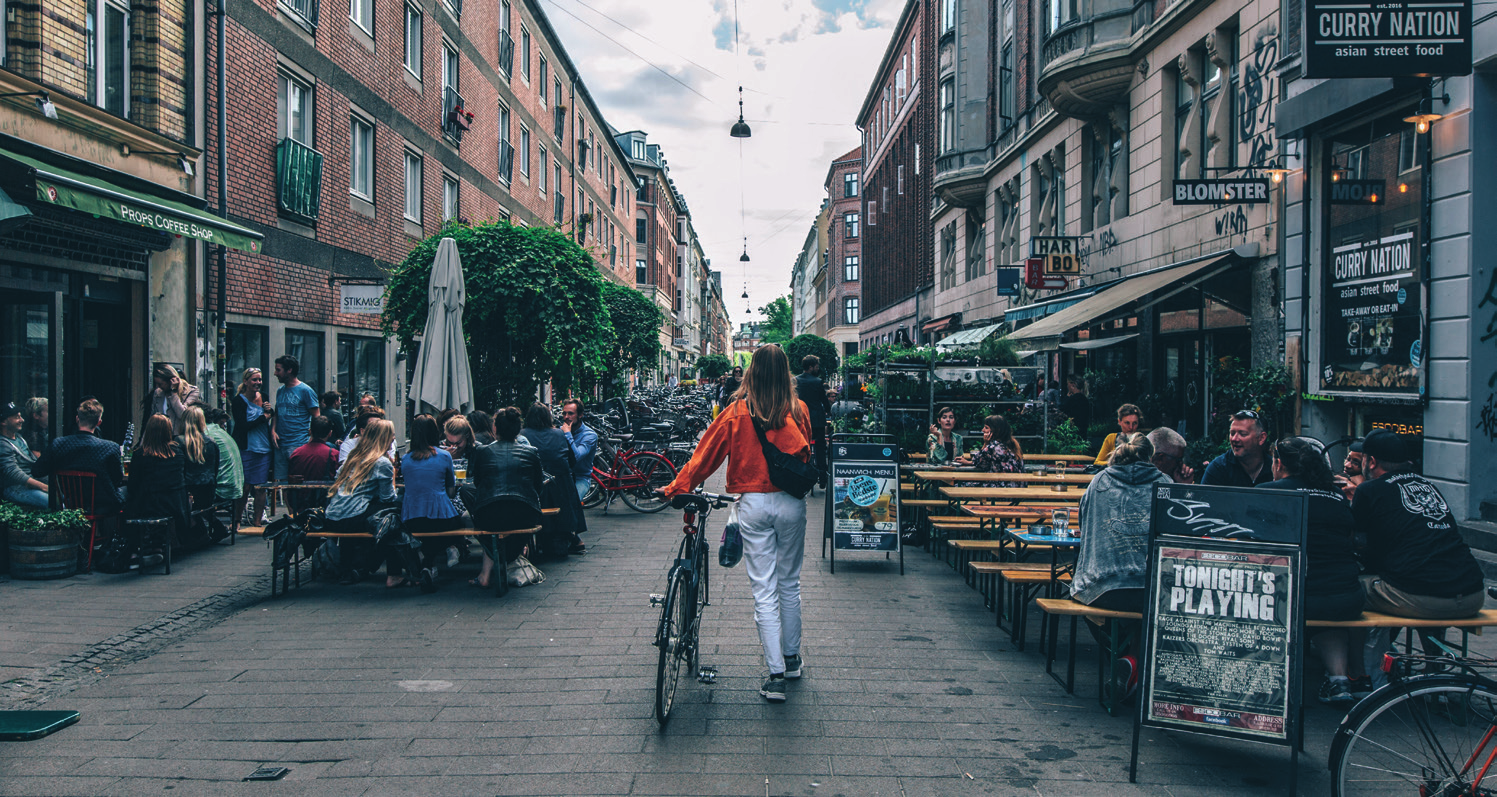 Smal gade i Nørrebro med caféer og restauranter på begge sider, hvor en kvinde trækker sin cykel gennem byens liv.