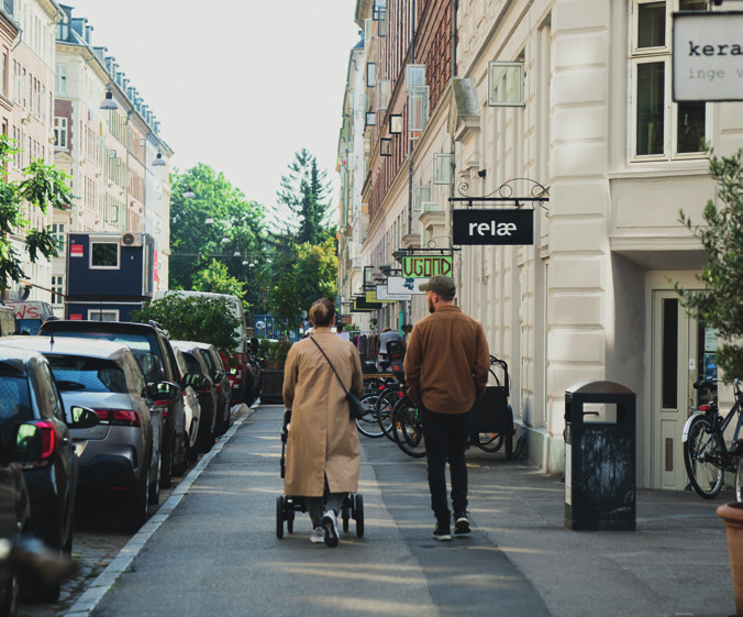 Et par med barnevogn går gennem Nørrebros gader omgivet af biler og byliv, set bagfra på en solrig dag.