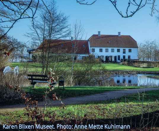 Karen Blixen Museum seen from a path with a bench and calm water.