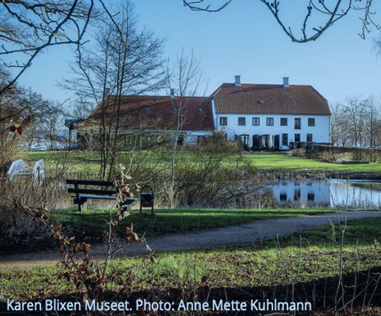 Karen Blixen Museum seen from a path with a bench and calm water.